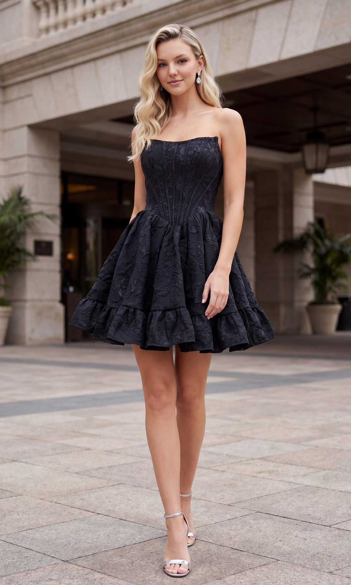A woman in a black Nox Anabel A7019 Strapless Floral Mini Hoco Dress with a corset-style bodice and heels stands outdoors on a paved area near columned architecture.